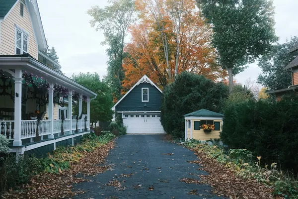 A paved driveway leading to a white garage door, clear of leaves down the middle, with autumn trees behind